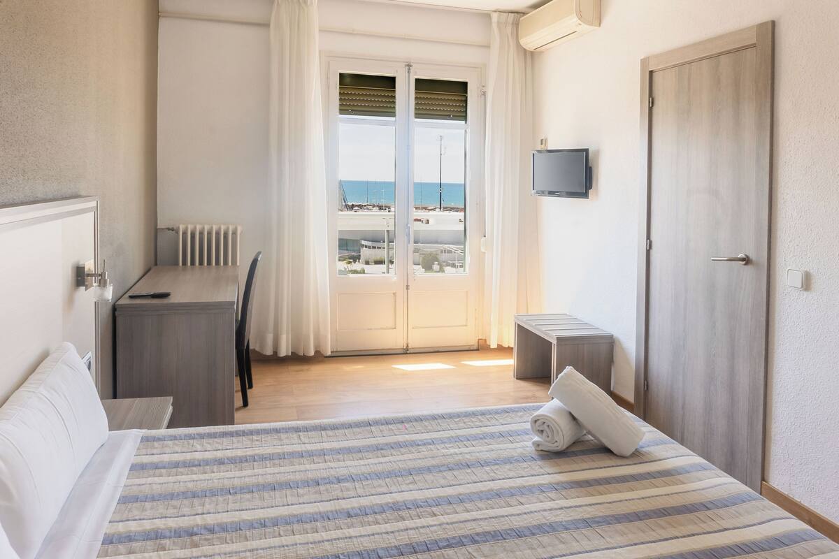 Habitación de hotel con cama, escritorio, silla, televisor montado en la pared y vista al mar por la ventana.