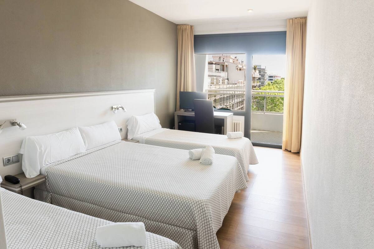 Habitación de hotel con dos camas, un escritorio y vistas a edificios a través de una ventana.