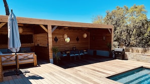 Een houten terras met een zwembad, zitplaatsen buiten en een barbecuehoek.