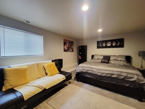A bedroom with a bed, a sofa, a window with blinds, a wall-mounted picture, and a ceiling light.