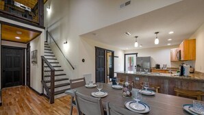 Un comedor espacioso con una mesa de madera dispuesta para cuatro comensales, una escalera que sube a un nivel superior y una cocina con armarios de madera y encimera.