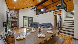 Amplia sala de estar con techo de madera, chimenea, una mesa de comedor para cuatro personas y una escalera que lleva a un nivel superior.