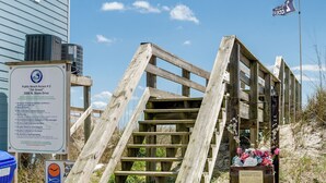 Eine Holztreppe führt zu einem Zugang zur Strandpromenade, mit einem Schild, das Strandregeln aufzeigt, und einer Gedenktafel.