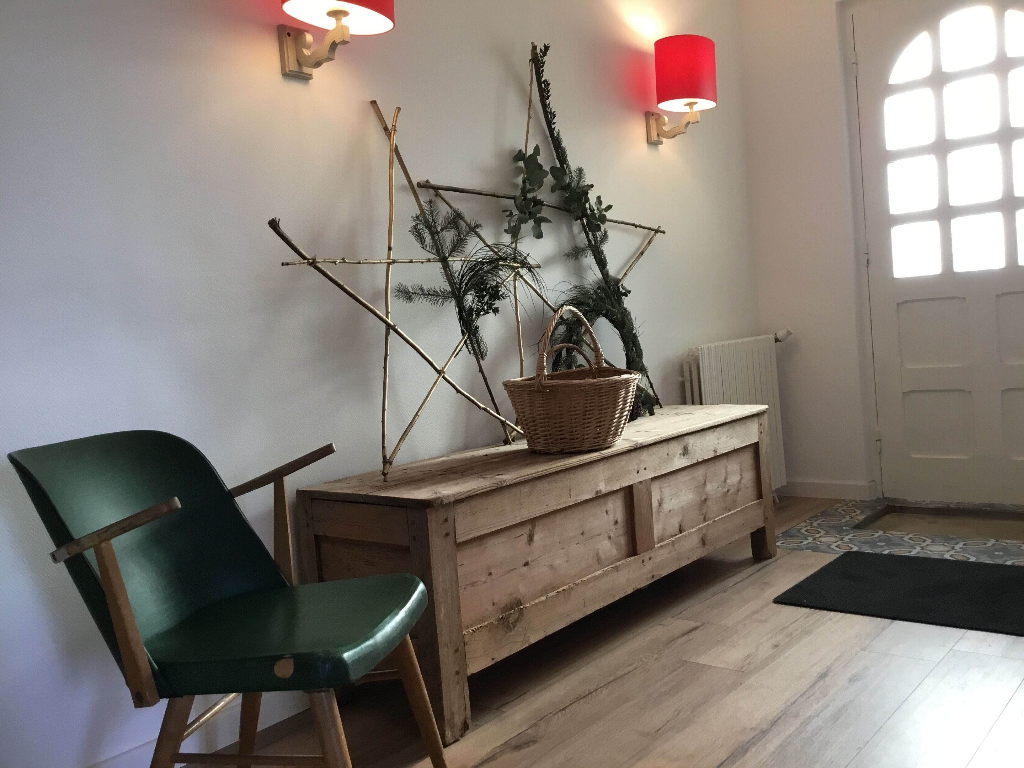 A room with a wooden bench, a green chair, a wall-mounted lamp, and a decorative plant arrangement.