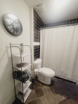 A bathroom with a toilet, a shelf with towels, a shower curtain, and a clock on the wall.