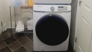 Two stacked Samsung washing machines in a laundry room with a metal shelf holding cleaning supplies.