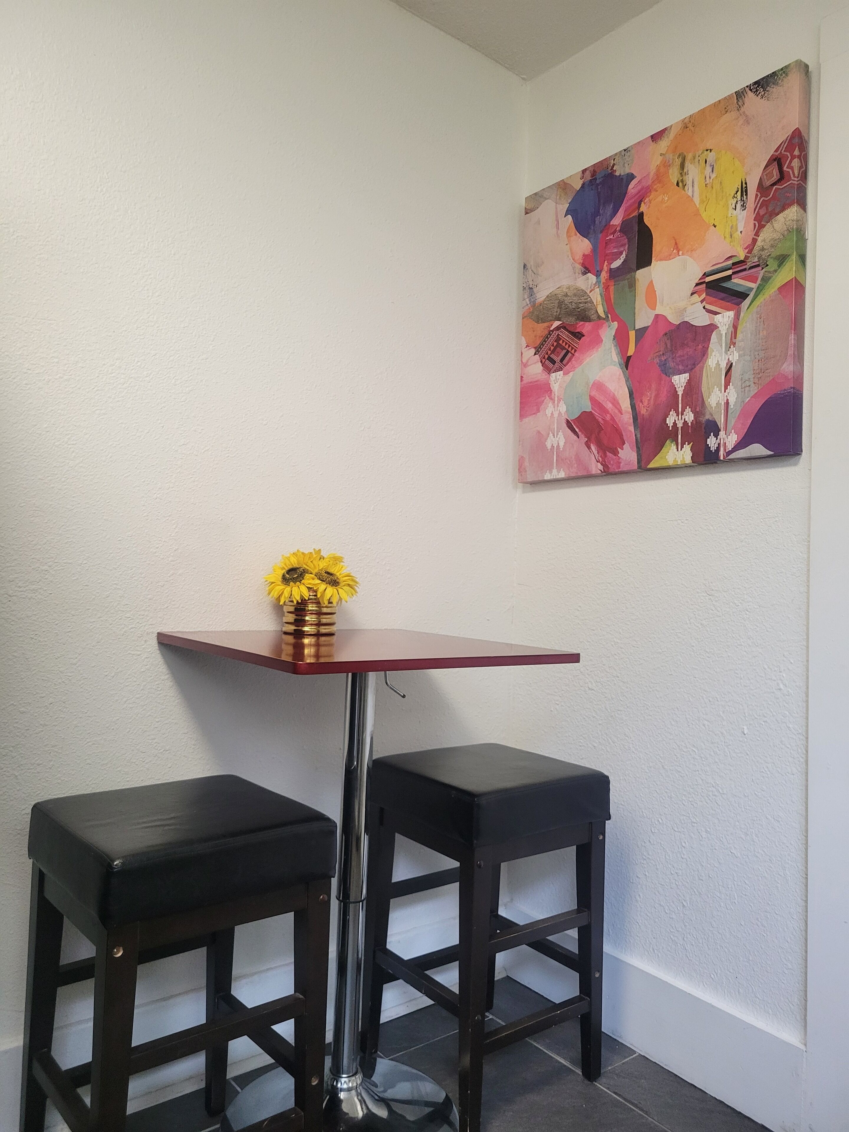 Una stanza d'angolo con un tavolino, due sgabelli neri, un vaso con fiori gialli e un quadro astratto dai colori vivaci appeso al muro.
