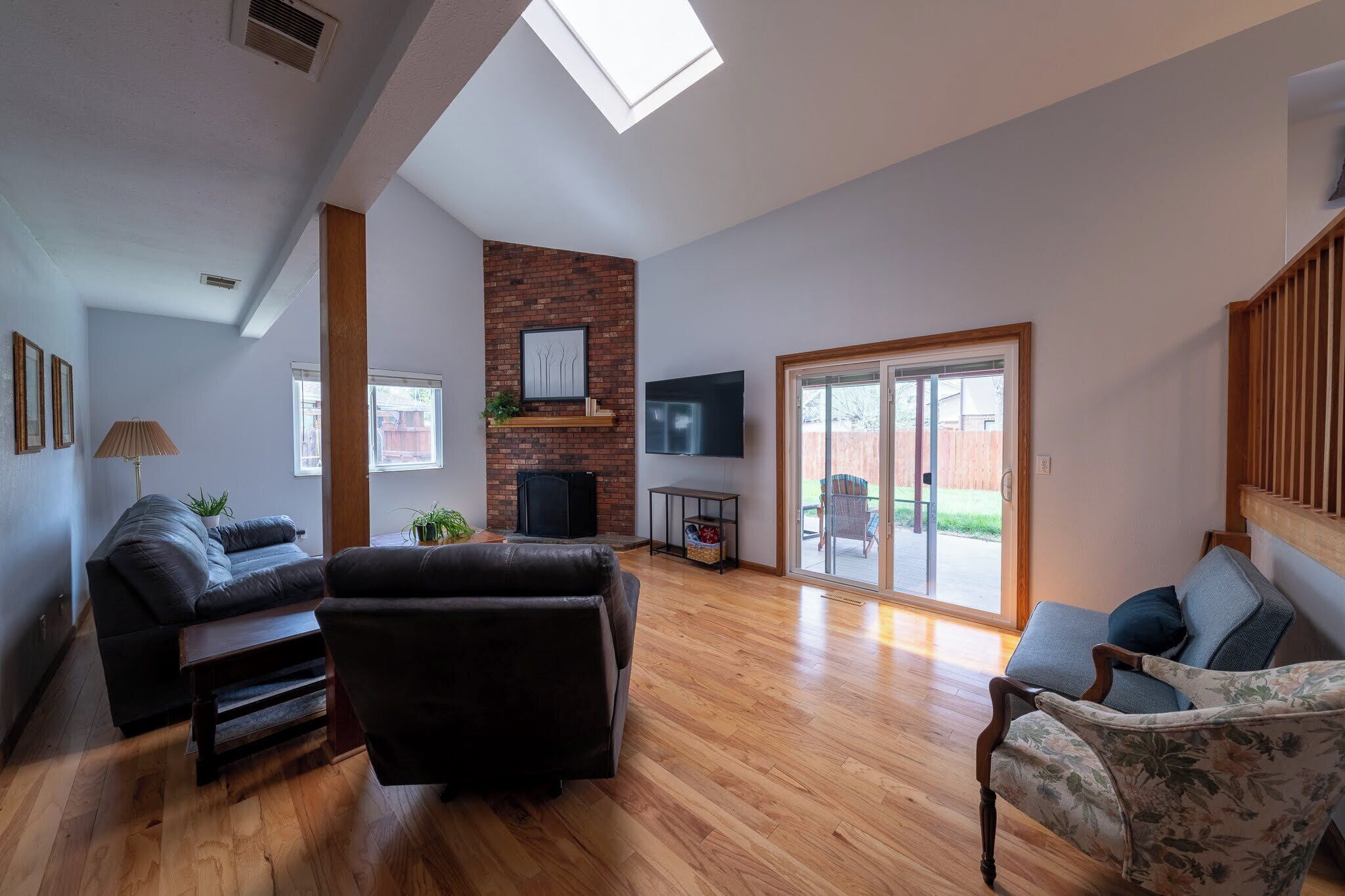 A spacious living room with a wooden floor, a brick accent wall, a flat-screen TV, and a sliding glass door leading to an outdoor area.