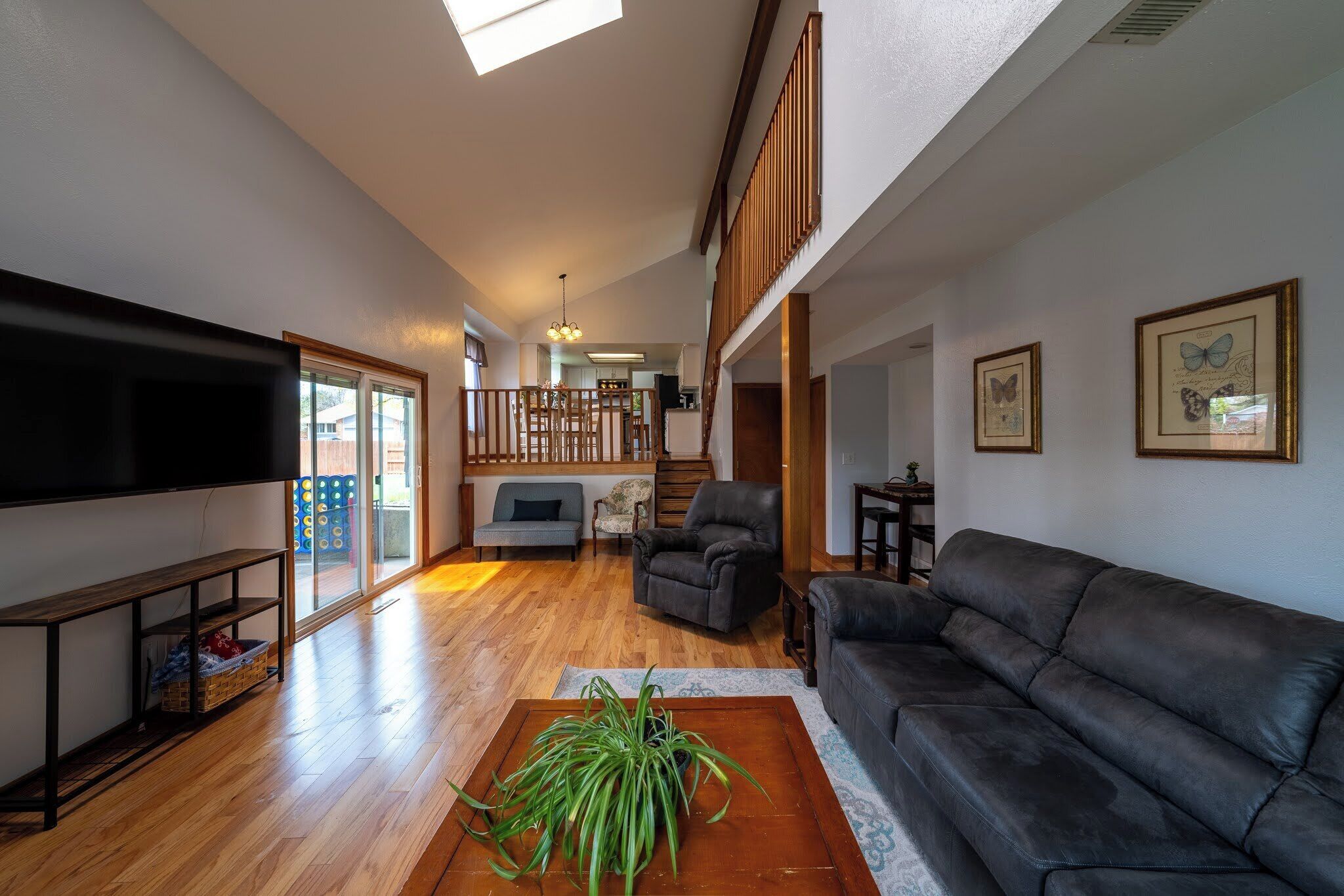 A spacious living room with a large flat-screen TV, a leather sofa, a coffee table with a plant, and a staircase leading to an upper level.