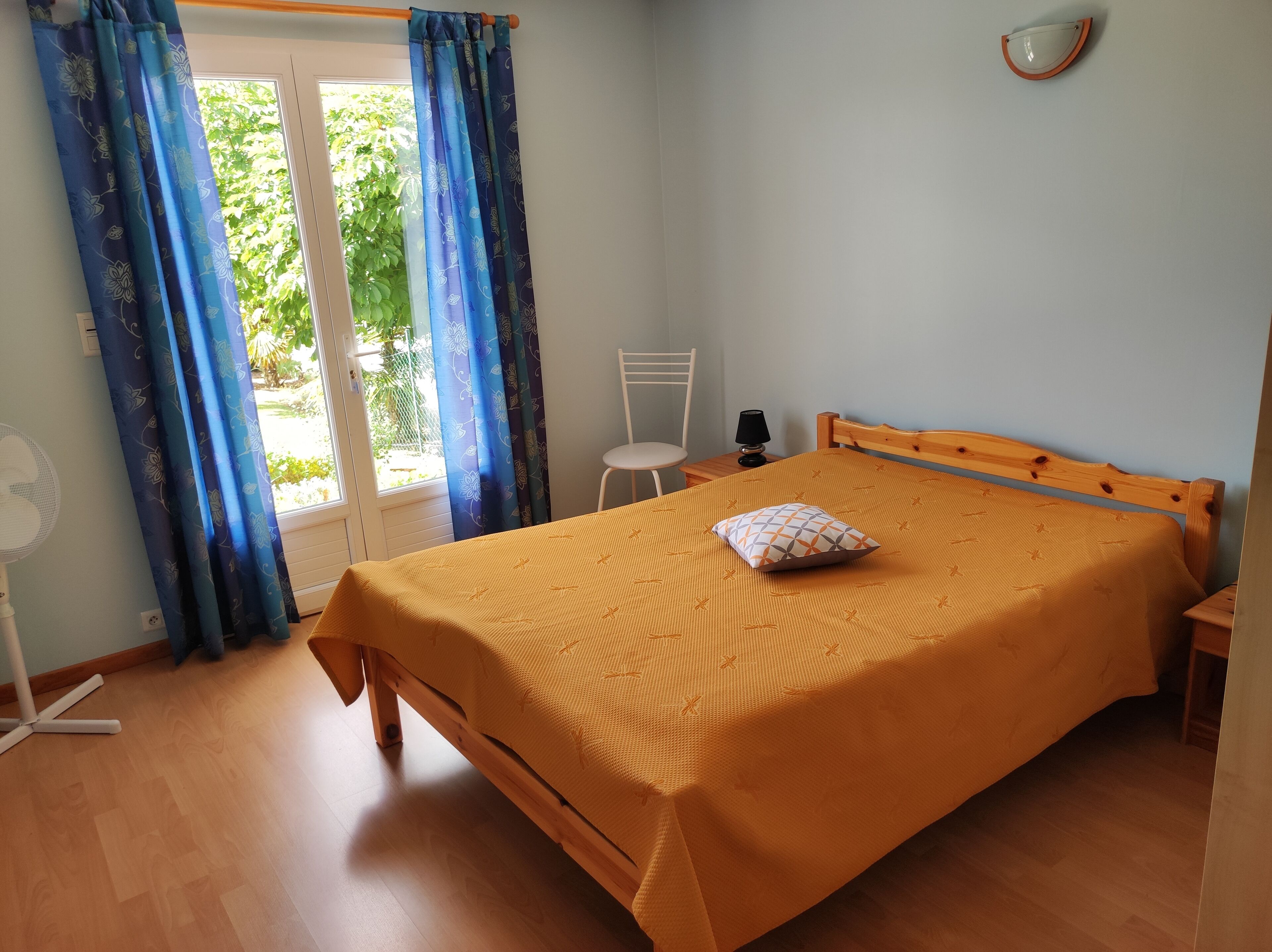 Ein Schlafzimmer mit Bett, Sessel, Nachttisch, Ventilator und einem Fenster mit Vorhängen.