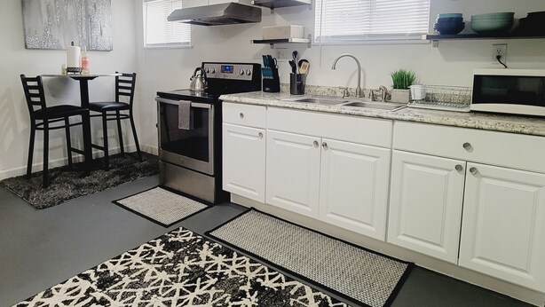 Una cocina moderna con armarios blancos, una estufa, un fregadero y un microondas. Hay dos sillas y una mesa pequeña con un tapete.