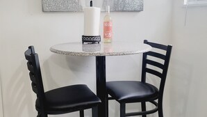Un comedor con una mesa redonda, dos sillas negras, un dispensador de toallas de papel blancas, una botella de vino y un cuadro abstracto plateado en la pared.