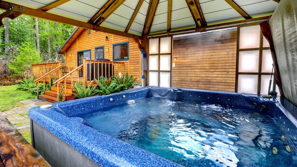 A hot tub under a wooden deck with a covered area, surrounded by a wooden cabin with a garage.