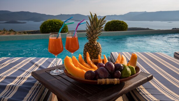 Una mesa con una bandeja de frutas y dos vasos de jugo de naranja, con vista a una piscina y una panorámica de montañas y agua.