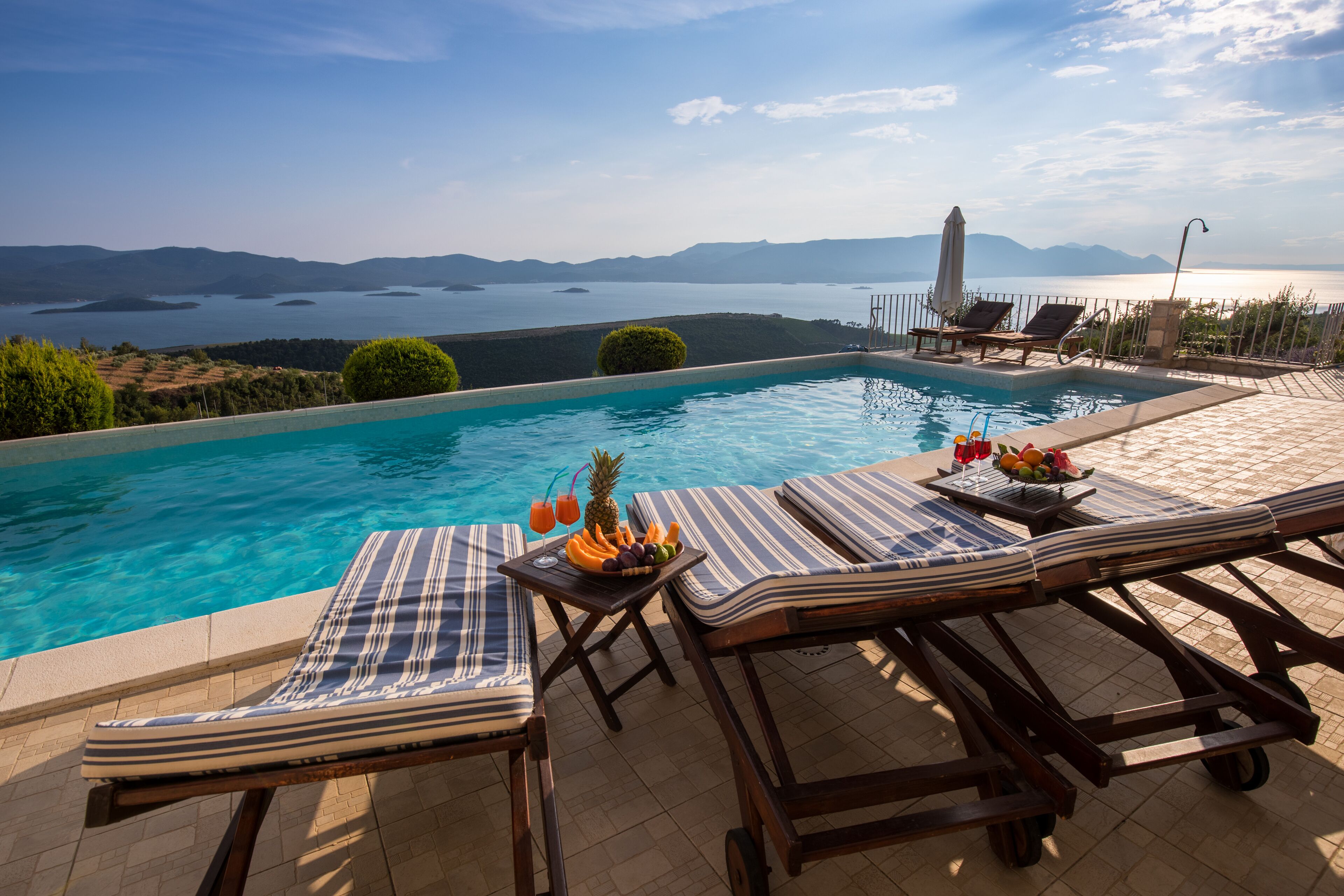 A poolside area with lounge chairs, a table with fruit and drinks, and a view of a lake and mountains.