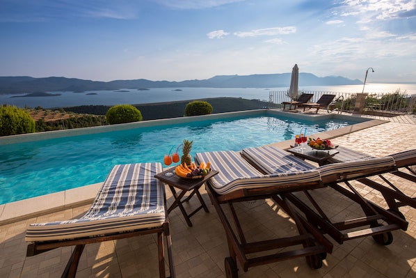 A poolside area with lounge chairs, a table with fruit and drinks, and a view of a lake and mountains.