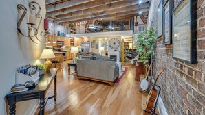 A spacious loft-style living area with exposed wooden beams, a mezzanine level, and a brick wall adorned with framed sheet music. - MtnMusicLoft,Find Your Rhythm in Heart of Downtown (Bristol)