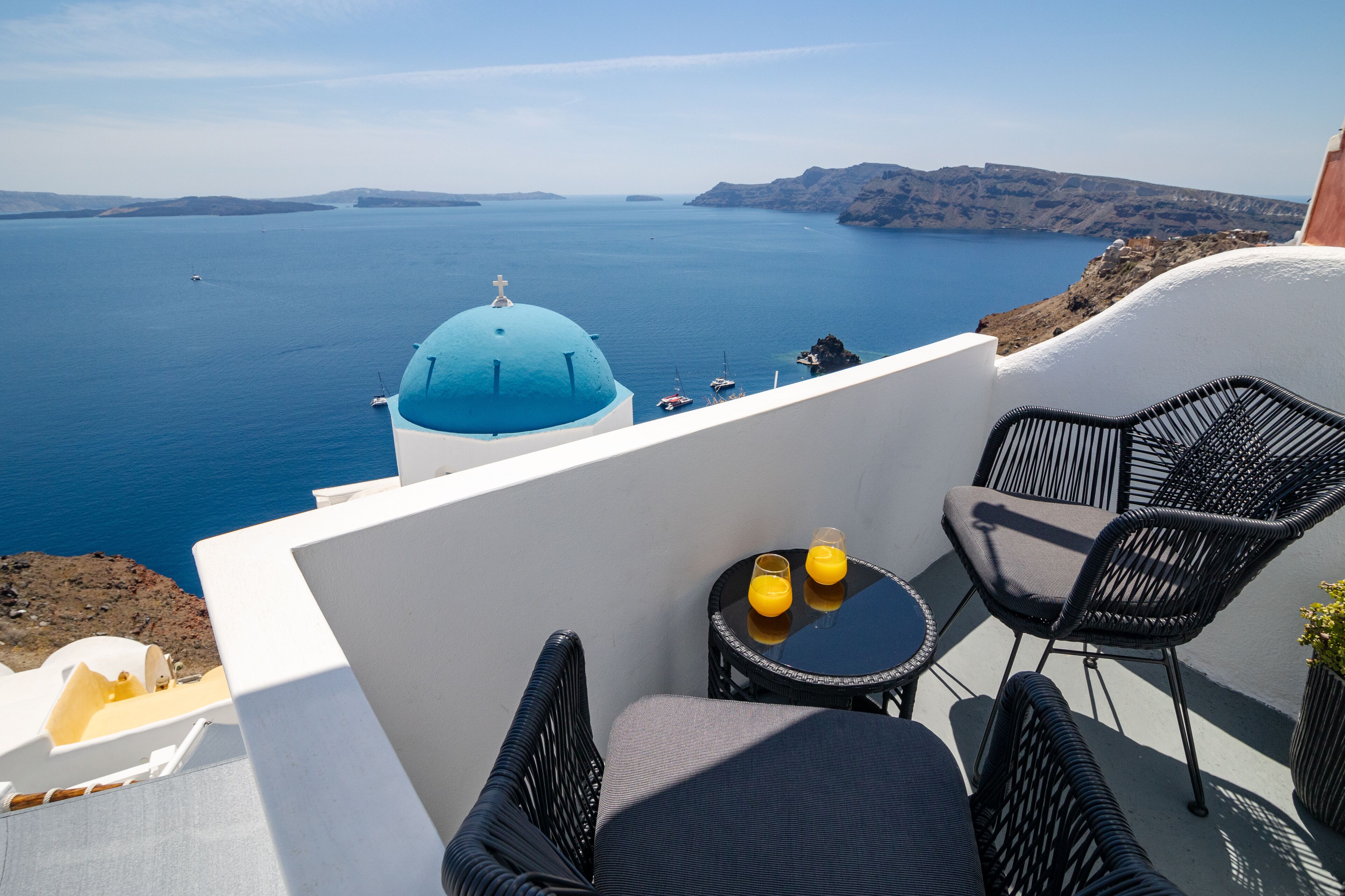A balcony with a view of the sea and a blue-domed building. There are two wicker chairs and a small round table with two glasses of orange juice.