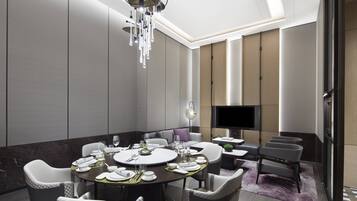 Uma sala de jantar moderna com uma mesa redonda posta para seis pessoas, rodeada por cadeiras estofadas. Um candelabro pendurado no teto e uma televisão de tela plana na parede.