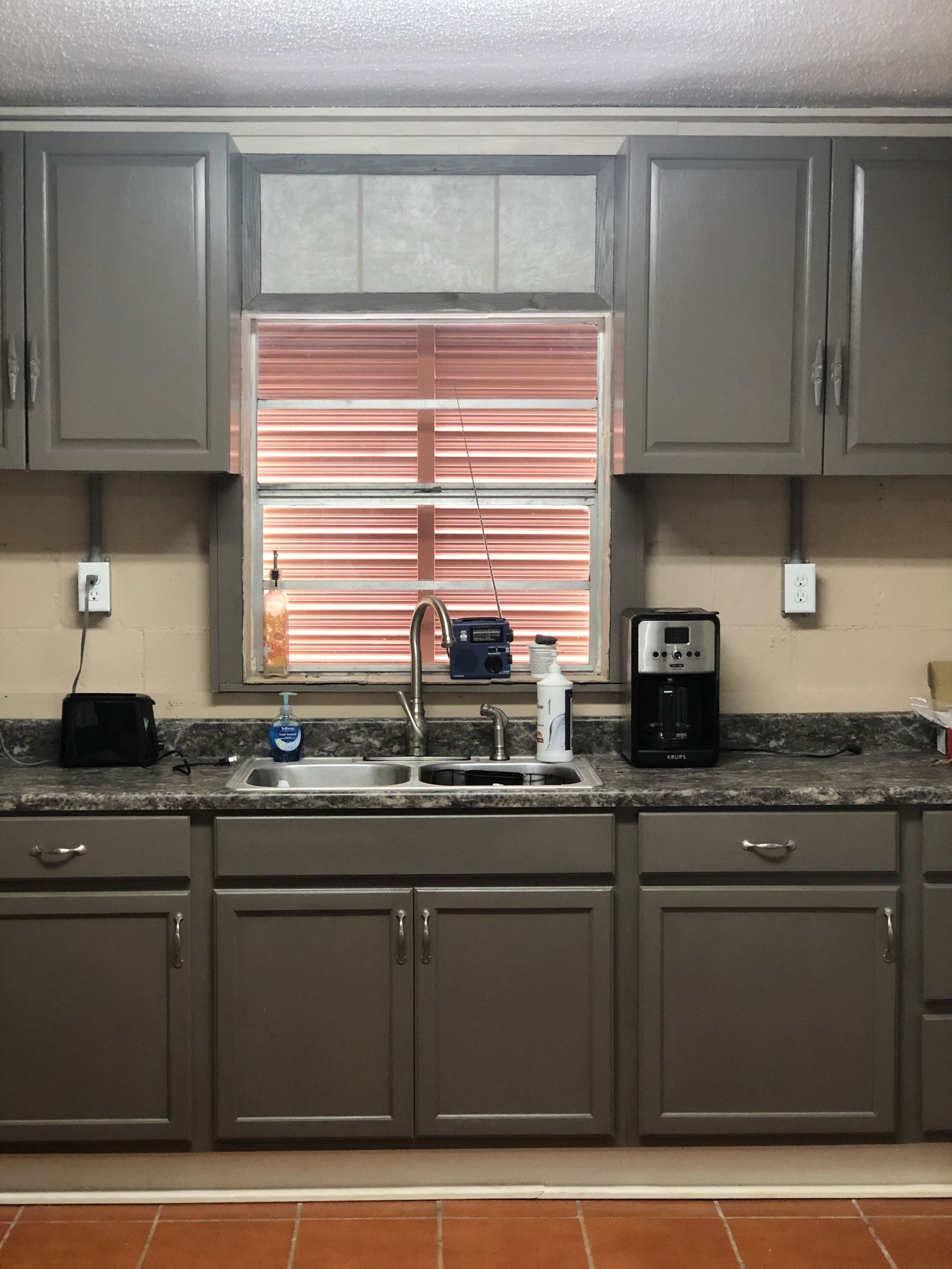 A kitchen with grey cabinets, a double sink, a coffee maker, and a window with blinds.