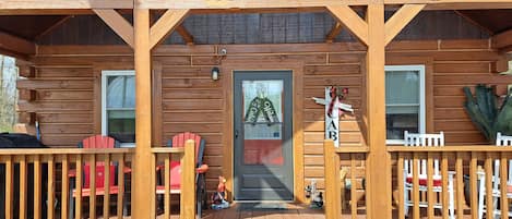 A wooden cabin with a porch, two windows, a door with a 'Welcome' sign, and a small outdoor seating area with chairs and a table.