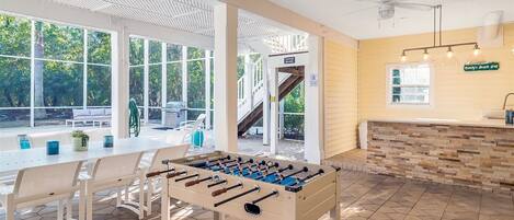 Amplo espaço interno com mesa de futebol, área de estar com mesa e cadeiras, e vista para uma área externa coberta com escada.