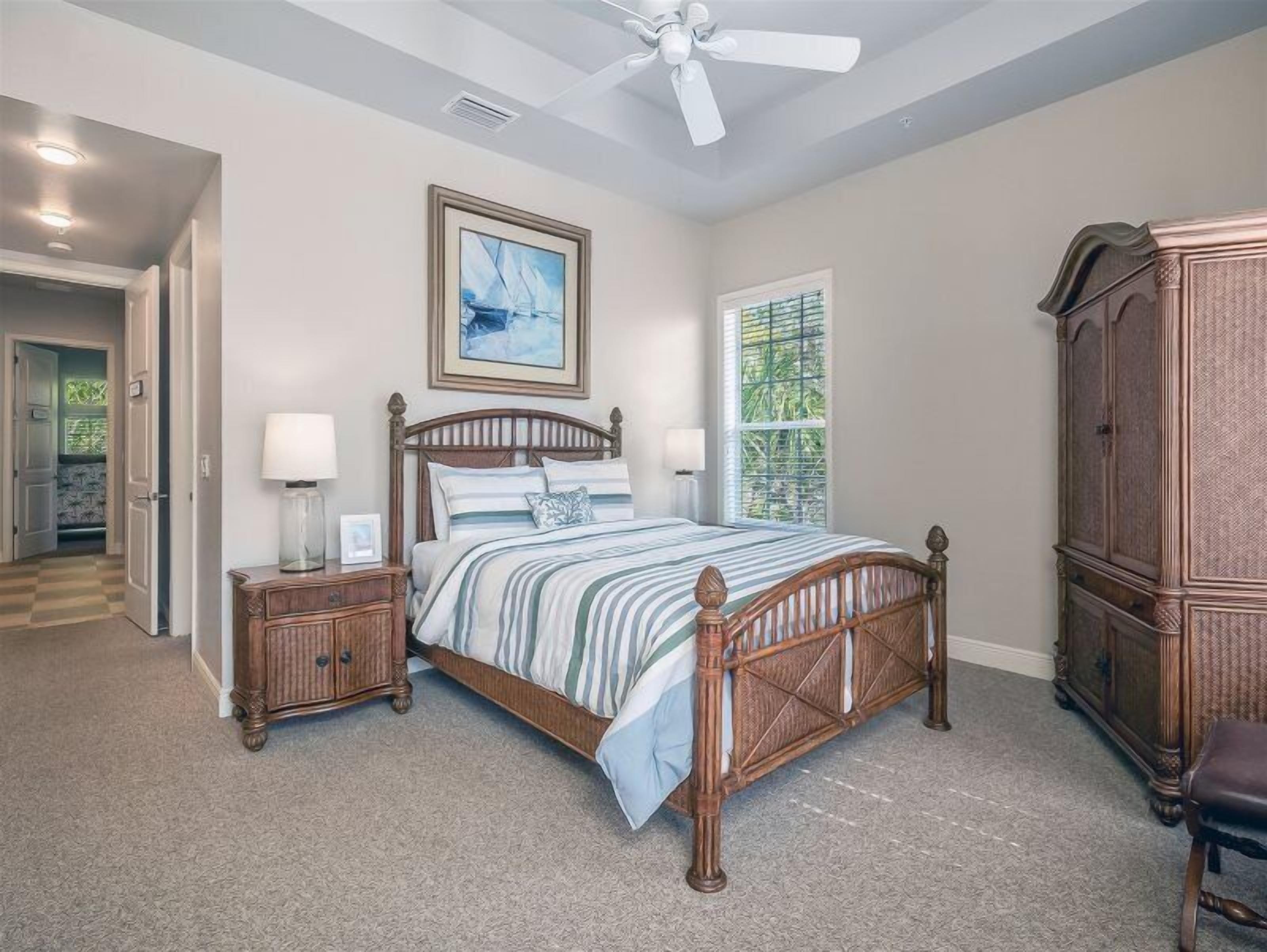 A bedroom with a wooden bed, nightstands, a ceiling fan, a window with a view of greenery, and a large wooden cabinet.