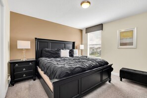 A bedroom with a black bed, two bedside tables with lamps, a window with blinds, and a framed picture on the wall.