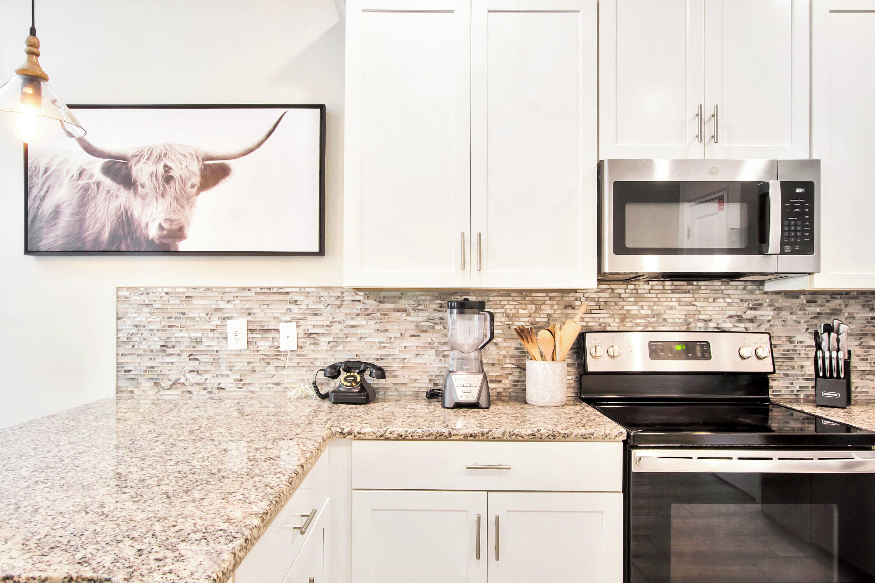 A modern kitchen with white cabinets, a granite countertop, a microwave, a stove, a blender, and a vintage telephone.