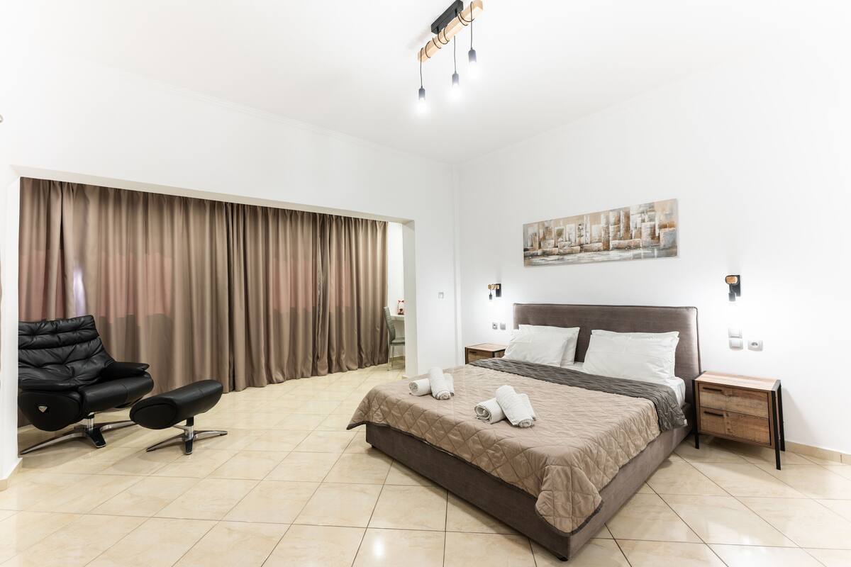A modern hotel room with a large bed, a black leather chair, a wooden nightstand, and a large window with curtains.