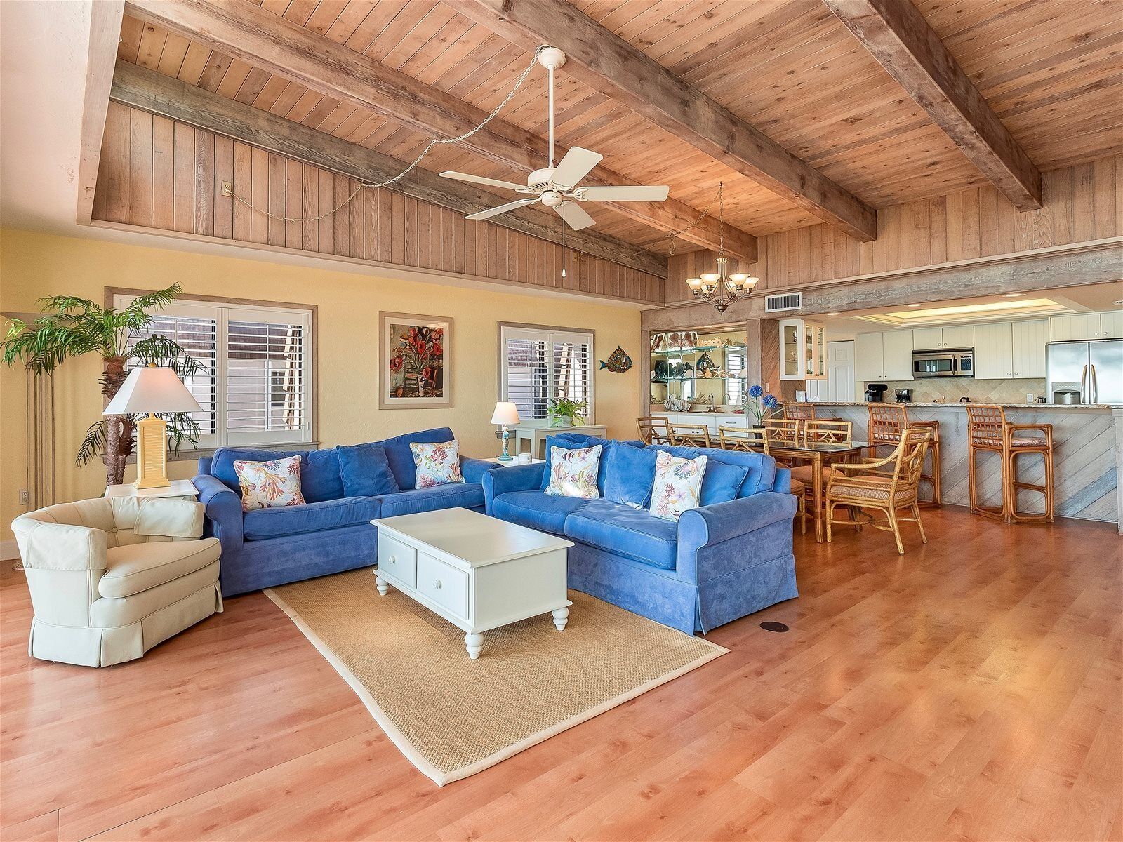 A spacious living area with a blue sectional sofa, a white coffee table, and a beige armchair. A kitchen with wooden cabinets and a dining area with wicker chairs is visible in the background.