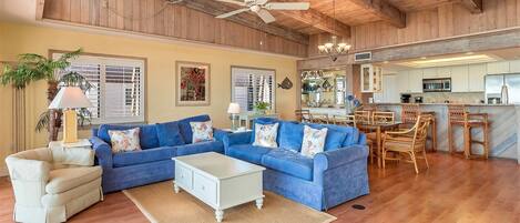 A spacious living area with a blue sectional sofa, a white coffee table, and a beige armchair. A kitchen with wooden cabinets and a dining area with wicker chairs is visible in the background.