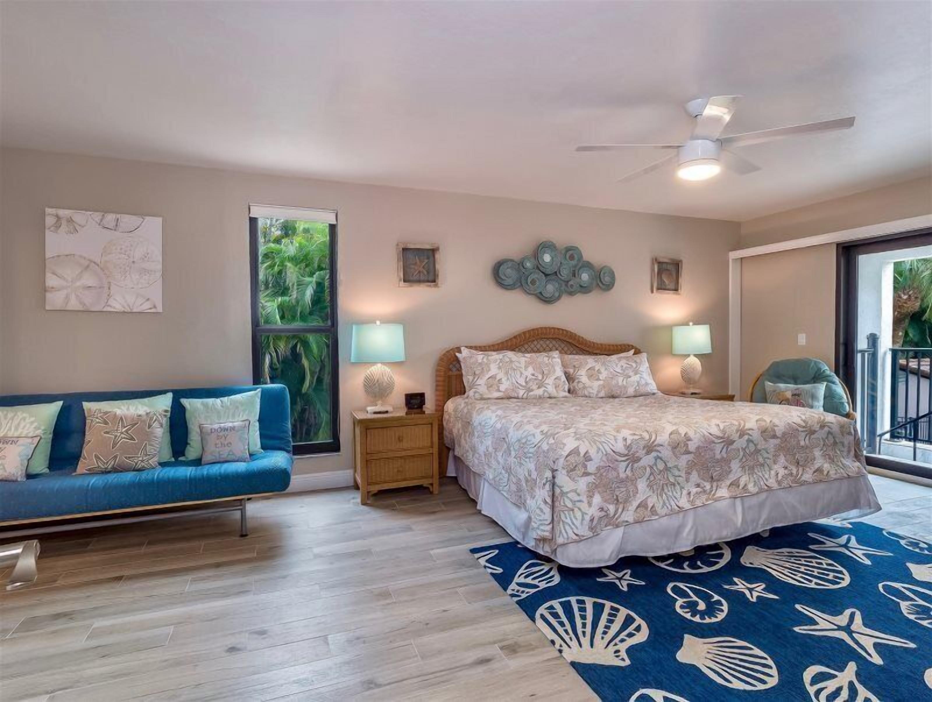 A bedroom with a bed, a blue sofa, a nightstand, a ceiling fan, and a large window with a view of greenery.