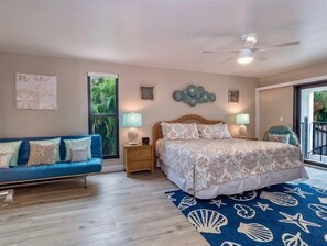 A bedroom with a bed, a blue sofa, a nightstand, a ceiling fan, and a large window with a view of greenery. - Shorewood Condos by Dream Vacation Rentals (Sanibel)