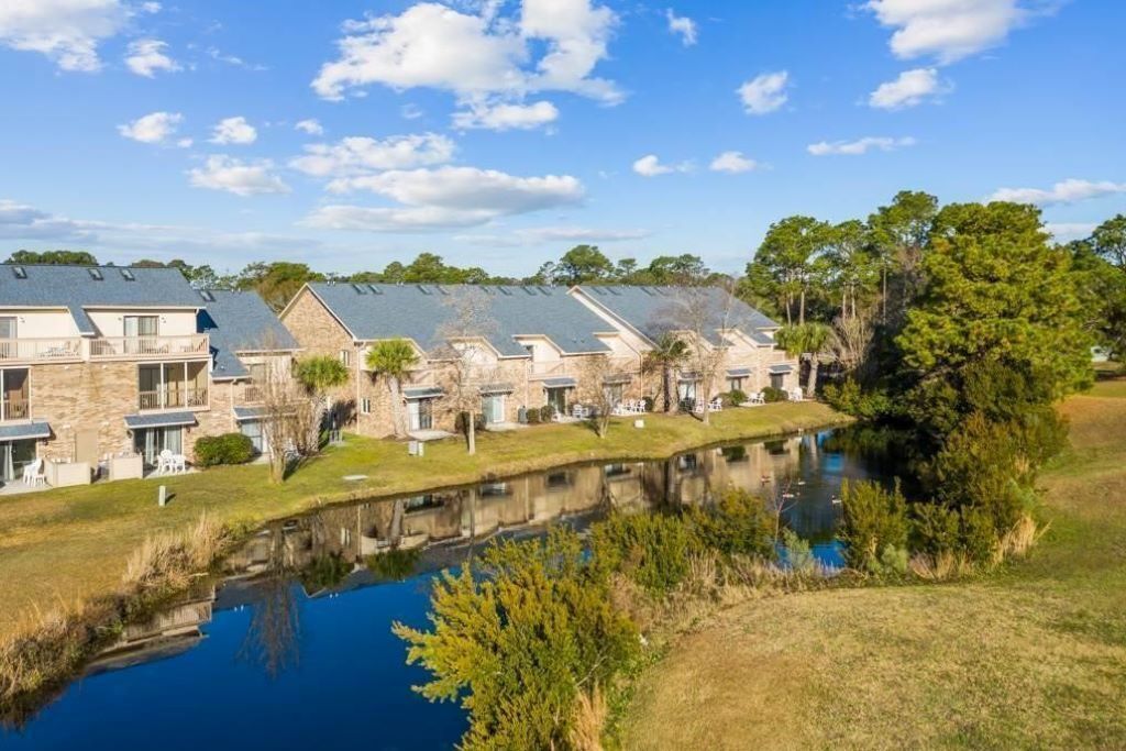 A row of stone buildings with balconies, a pond in the foreground, and trees in the background.