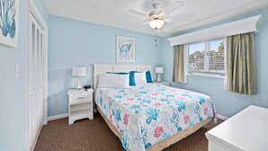 Une chambre à coucher comprenant un lit, des tables de chevet, un ventilateur de plafond, une fenêtre avec des rideaux et des œuvres d’art accrochées aux murs.