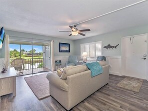 A living room with a sofa, a desk, and a ceiling fan. There is a door leading to a balcony with a view of a golf course. - Spanish Cay by Dream Vacation Rentals (Sanibel)