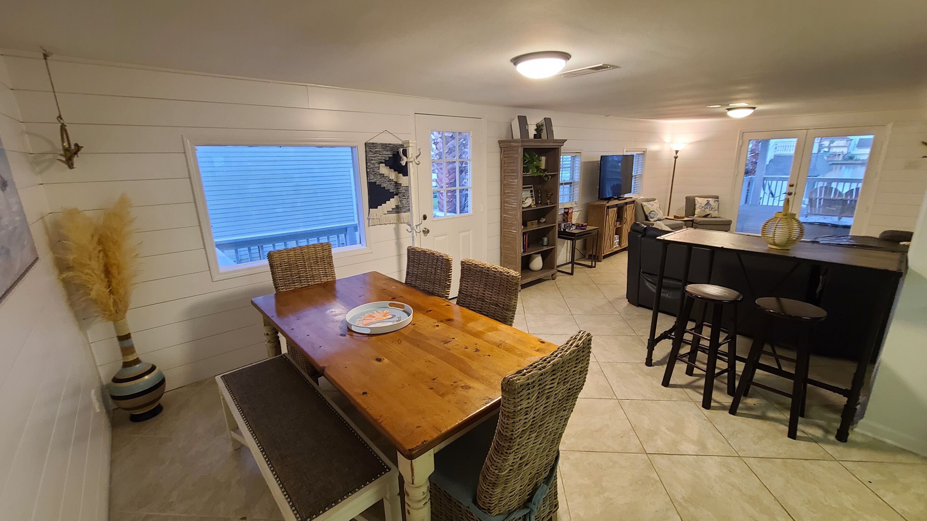 A dining area with a wooden table and wicker chairs, a bookshelf, and a bar area with stools.