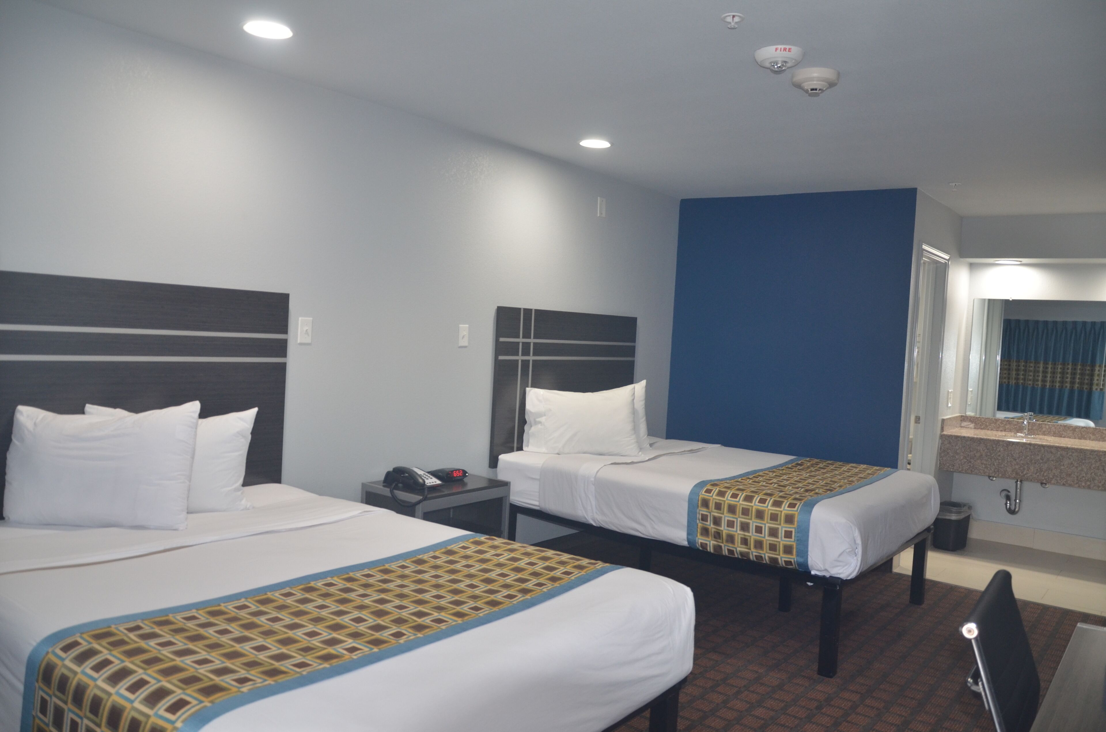A hotel room with two beds, a desk, a chair, a telephone, and a bathroom with a mirror and sink.