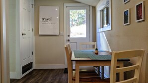 A room with a wooden floor, a white door, a framed word 'travel' on the wall, a window with a view of trees, and a table with chairs. - Traveler's Mountain Retreat w/ Grotto Cave Room (Idaho Springs)