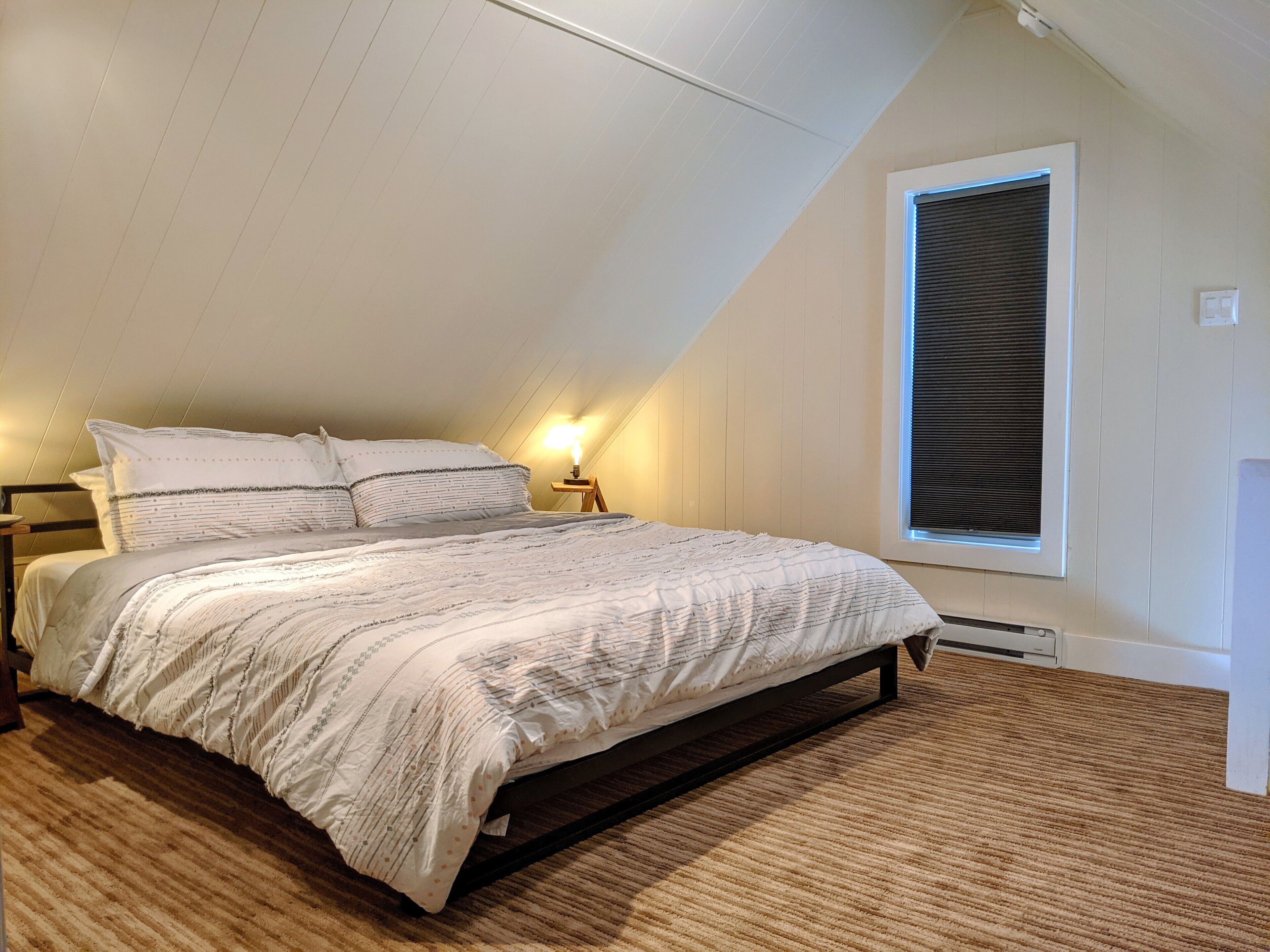 Ein Schlafzimmer mit einem Bett, einem Nachttisch, einer Lampe, einem Fenster mit Jalousien und einer Heizung.