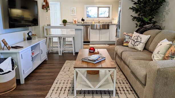 A modern living room with a sofa, coffee table, and a TV. There is a kitchen in the background with white cabinets and a stainless steel fridge. - Traveler's Mountain Retreat w/ Grotto Cave Room (Idaho Springs)