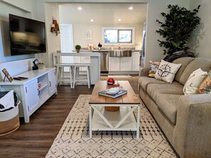 A modern living room with a sofa, coffee table, and a TV. There is a kitchen in the background with white cabinets and a stainless steel fridge. - Traveler's Mountain Retreat w/ Grotto Cave Room (Idaho Springs)
