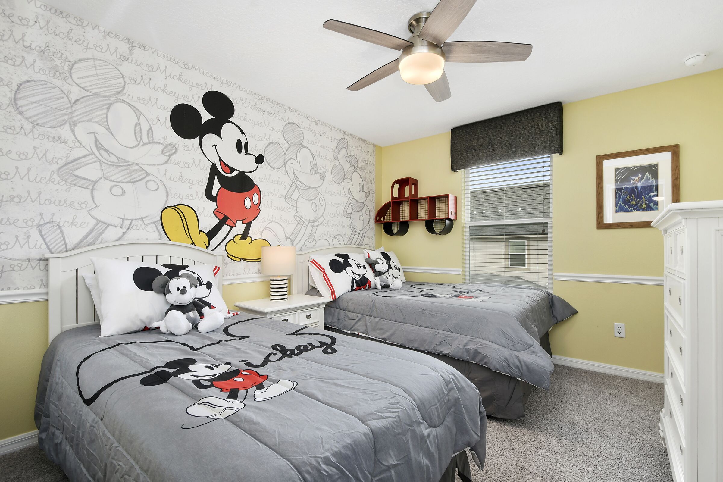 A bedroom with two beds, a Mickey Mouse mural, a ceiling fan, and a framed picture on the wall.