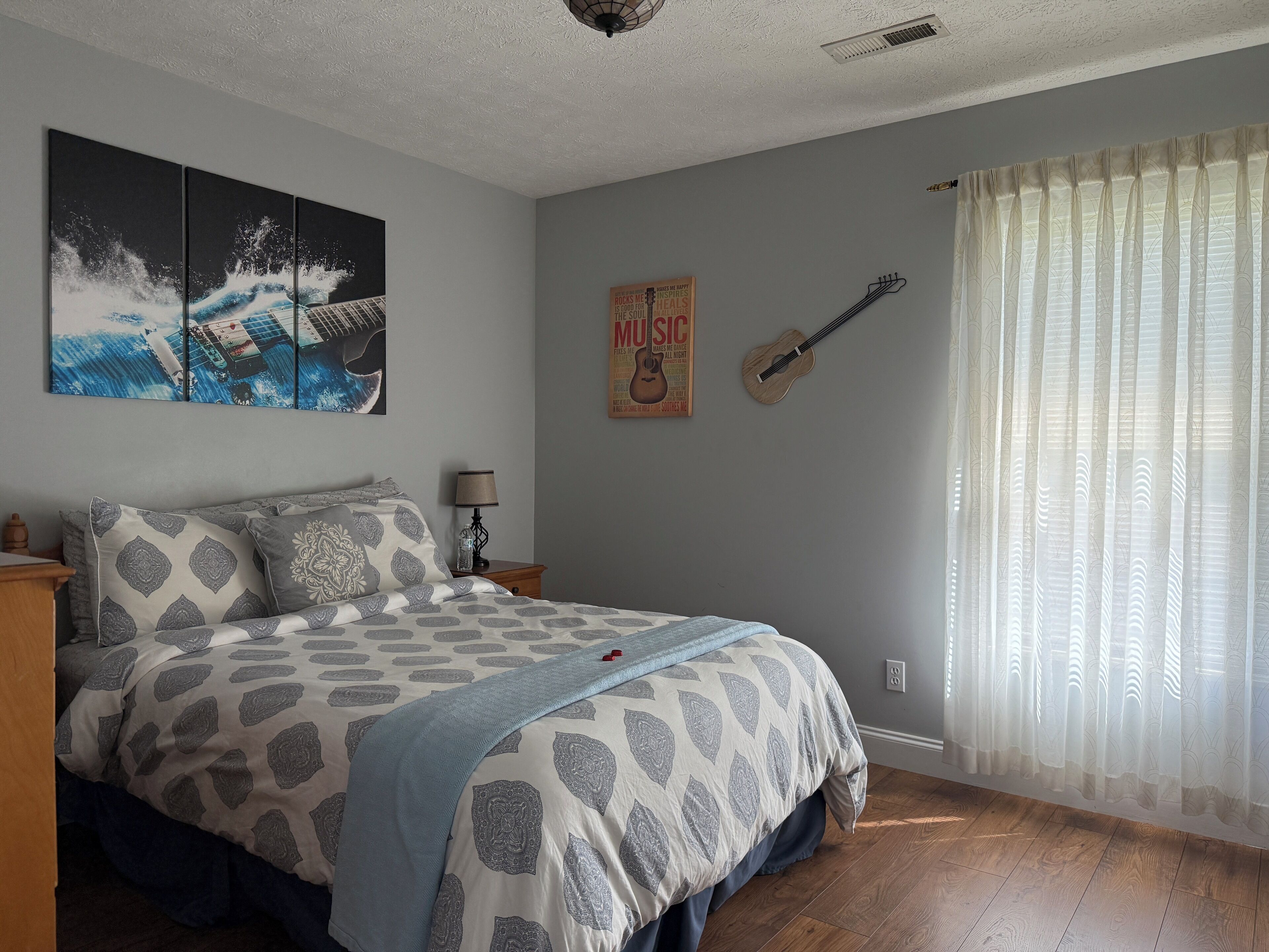 Een slaapkamer met een bed, een aan de muur bevestigde gitaar, een poster en een raam met dunne gordijnen.