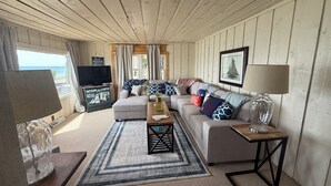 Una sala de estar con sofá, mesa de centro y televisor, paredes de madera y vista al océano.