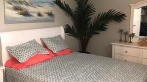 A bedroom with a bed, a ceiling fan, a large plant, a painting, and a dresser.