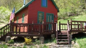 A red cabin with a green roof, a wooden deck, and a stone staircase leading up to the entrance.