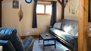 A cozy living room with a sofa, a coffee table, and a leather chair. There's a window with curtains, a wall clock, and a welcome sign.