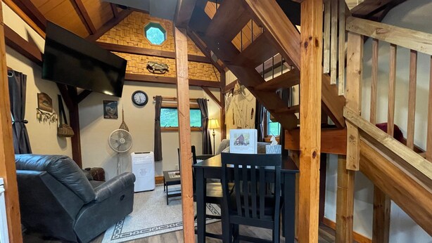 A cozy living area with a wooden ceiling, a black sofa, a dining table with chairs, and a small kitchen area with a refrigerator and a fan.